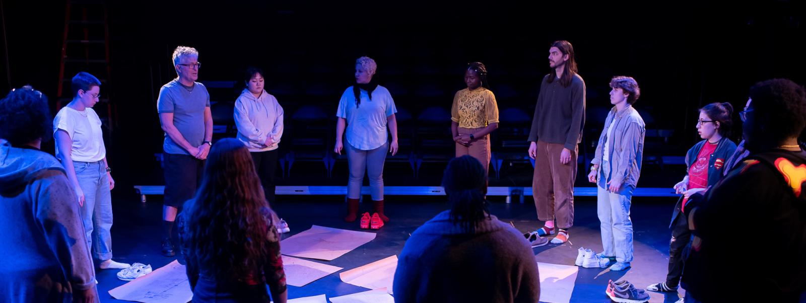 A diverse group of people stands in a dimly lit circle, surrounded by papers on the floor.