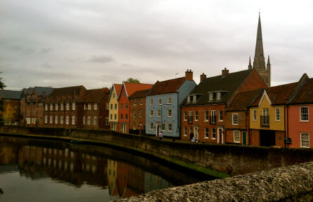 A row of colorful houses lines a calm riverside under an overcast sky, with a tall church spire in the background reflecting a quaint, serene atmosphere.
