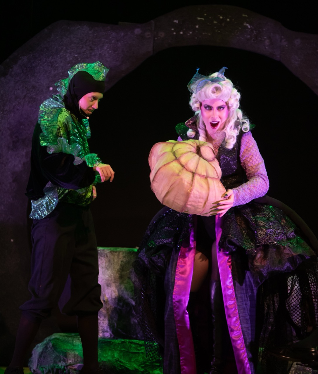 On a dark theatrical stage, a white-wigged performer cradles a giant pumpkin as a green-clad companion watches, creating a magical mood.