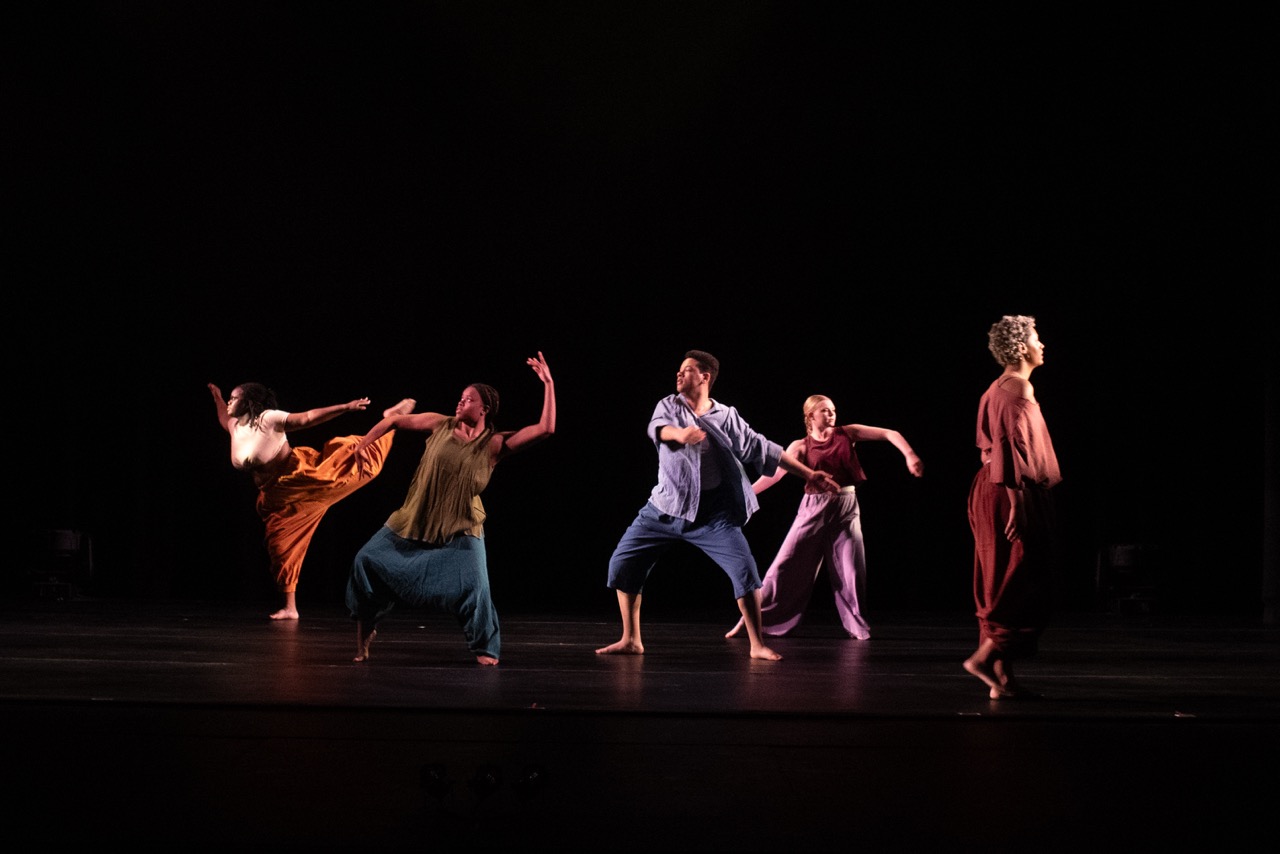 Contemporary dancers in colorful costumes perform expressive, energetic poses across a dark stage, creating a dramatic, dynamic mood.