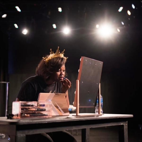 A woman wearing a decorative headpiece applies makeup at a table with a mirror on a dimly lit stage, conveying focus and anticipation.
