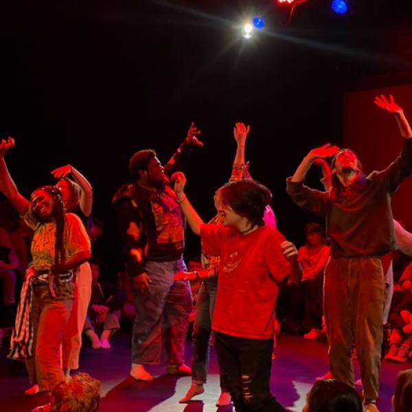 A lively group of people energetically dance under colorful stage lights, with onlookers seated around them. The atmosphere is joyful and dynamic.